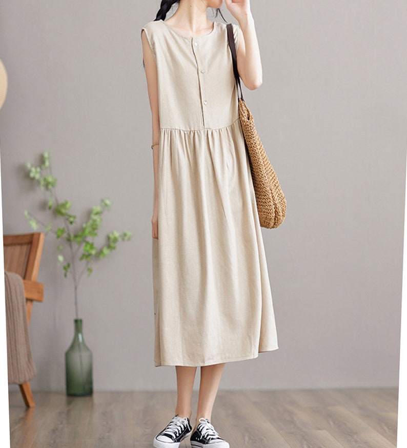 May include: A beige sleeveless linen dress with a button-up front and a gathered skirt. The dress is midi-length and has a relaxed fit.