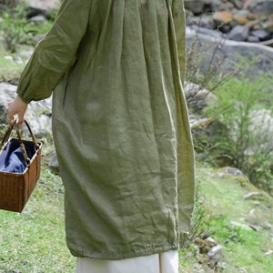 May include: A long, loose-fitting, green linen dress with a gathered neckline and a keyhole opening. The dress has long sleeves with gathered cuffs and a slightly gathered hem.