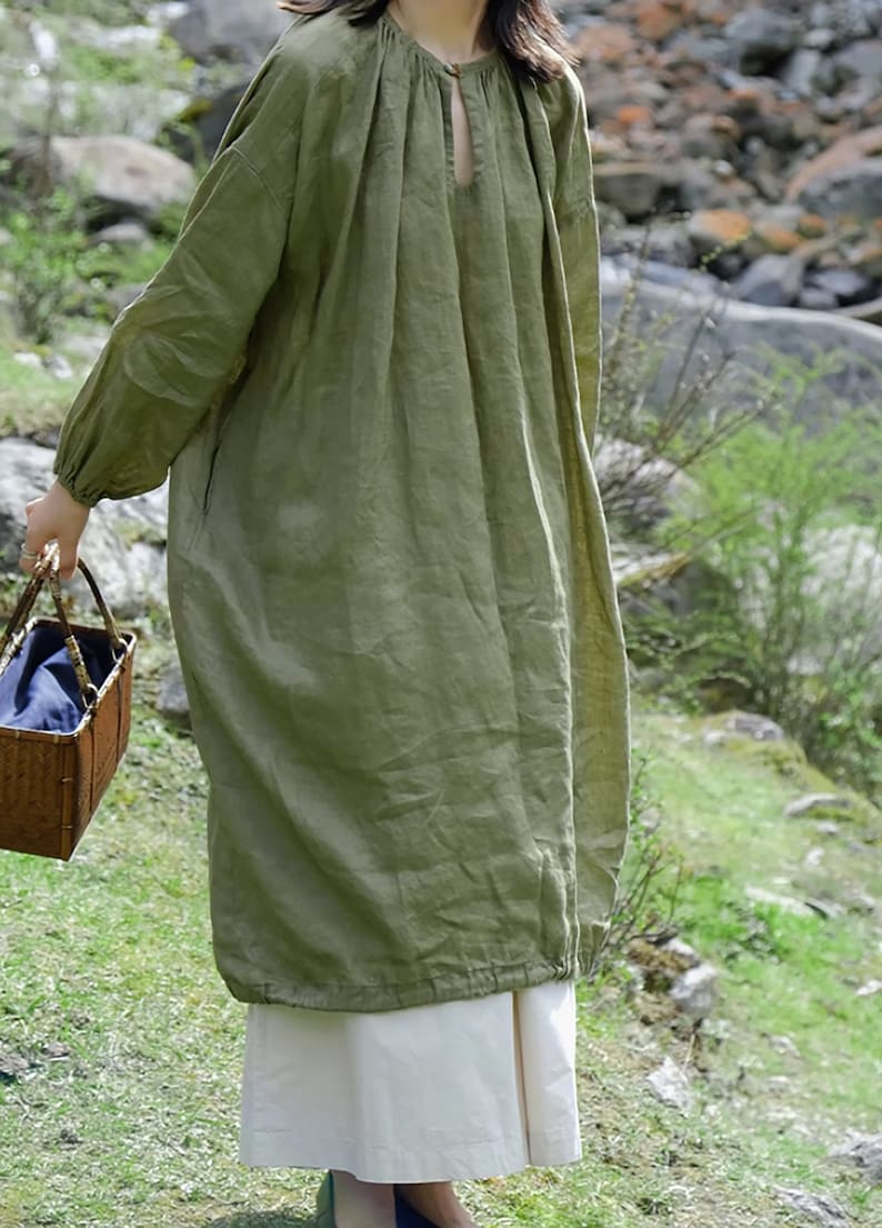 May include: A long, loose-fitting, green linen dress with a gathered neckline and a keyhole opening. The dress has long sleeves with gathered cuffs and a slightly gathered hem.