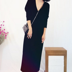 May include: A black velvet maxi dress with a v-neckline and long sleeves. The dress is worn by a person with short dark hair and red lipstick. The person is holding a small black purse with a silver chain strap.