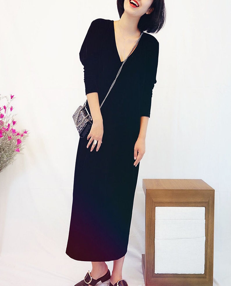 May include: A black velvet maxi dress with a v-neckline and long sleeves. The dress is worn by a person with short dark hair and red lipstick. The person is holding a small black purse with a silver chain strap.