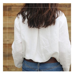 May include: A white linen shirt with long sleeves and a button-up front. The shirt is tucked into a pair of blue jeans and a brown belt is worn around the waist.