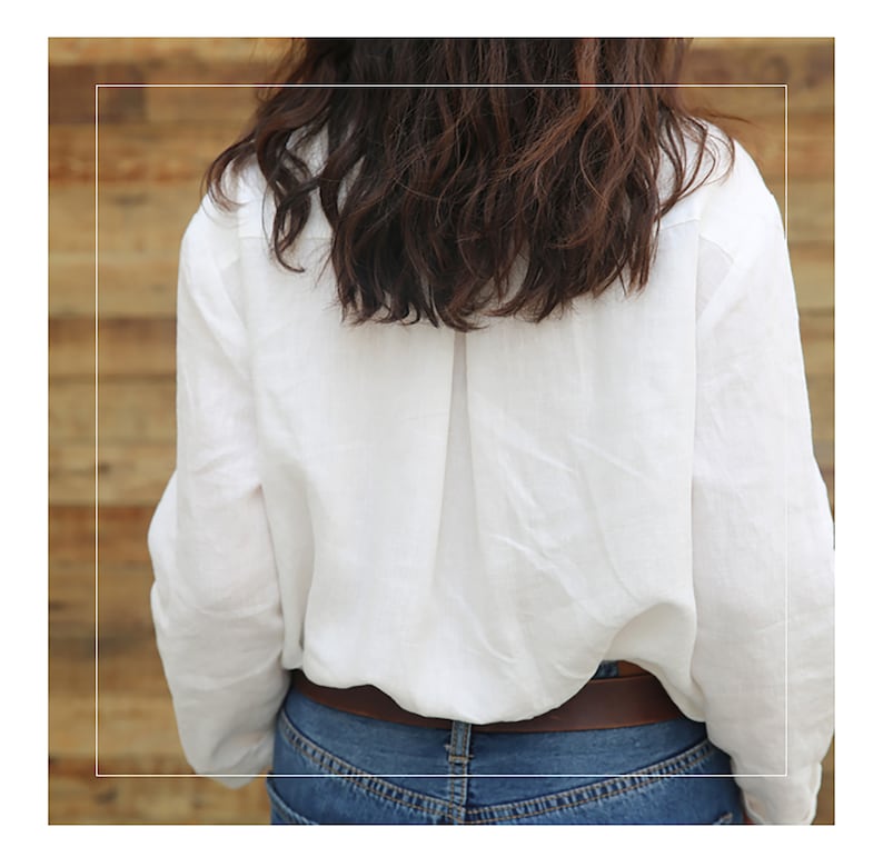 May include: A white linen shirt with long sleeves and a button-up front. The shirt is tucked into a pair of blue jeans and a brown belt is worn around the waist.