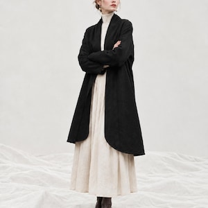 May include: A black open-front coat with long sleeves and a shawl collar, worn over a cream-coloured mock-neck top and a long, pleated cream-coloured skirt. The outfit is completed with dark brown ankle boots, creating a layered ensemble.