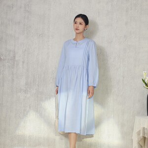 May include: A light blue long-sleeve dress with a round neckline and pleated skirt. The dress features a button detail at the neckline and is paired with brown sandals. A white purse hangs from the shoulder.