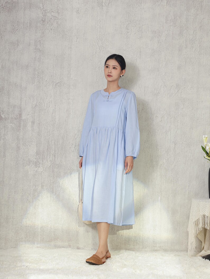 May include: A light blue long-sleeve dress with a round neckline and pleated skirt. The dress features a button detail at the neckline and is paired with brown sandals. A white purse hangs from the shoulder.
