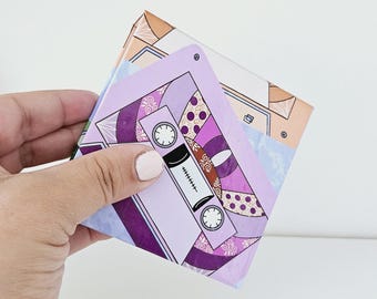 May include: A square coaster featuring a graphic illustration of a purple cassette tape with intricate patterns. The design includes shades of lavender, deep violet, and gold accents. The cassette is centered on a light blue and tan background.
