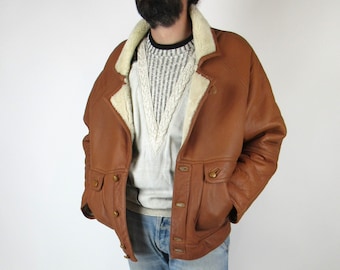 Op de afbeelding: Een bruin leren jack met een bontkraag en voering. Het jack heeft een knoopsluiting en twee klepzakken.