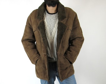 Op de afbeelding: Een bruine schapenvachtjas met een bontkraag en een knoopsluiting. De jas wordt gedragen over een witte trui en een blauwe jeans.