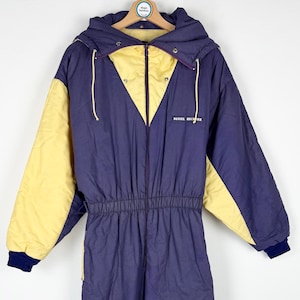 Könnte beinhalten: Ein Vintage-Ski-Anzug in Marineblau und Gelb mit Kapuze. Der Anzug hat einen Reißverschluss vorne, einen elastischen Bund und marineblaue Bündchen. Der Text "DANIEL RICHTER" ist auf der Brust aufgedruckt. Auf einem Holzbügel aufgehängt.
