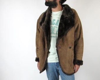 Op de afbeelding: Een bruin schapenvachten jack met een bontkraag en een knoopsluiting. Het jack wordt gedragen over een wit T-shirt met een grafische print. De persoon draagt een blauwe jeans en zwarte schoenen.