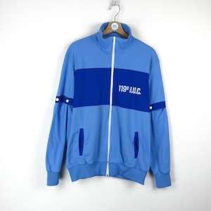 Peut inclure: Une veste zippée bleu clair avec une bande bleue sur la poitrine et le texte "119th A.U.C." en blanc. La veste a une fermeture éclair blanche et des finitions bleues sur les poches.