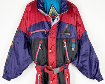 May include: A vintage ski suit in red, black, and purple. The one-piece suit features a high collar, multiple zippered pockets, and a belt. The design includes color-blocked panels and geometric accents, with a retro aesthetic. The suit is hanging on a wooden hanger.