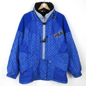 May include: A blue Fila winter jacket with a geometric pattern. The jacket has a black collar, a gray zipper, and the Fila logo on the chest. It features pockets and a hood.