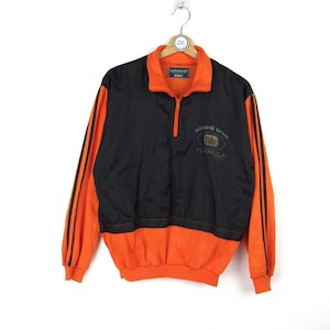 Può includere: Giacca con zip nera e arancione con strisce bianche e nere sulle maniche. La giacca ha un logo "Minardi Team Formula 1" sul davanti.