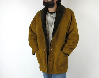 Op de afbeelding: Een bruine suède shearling jas met een grote kraag. De jas heeft een knoopsluiting en twee zakken. De persoon die de jas draagt, draagt ook een blauwe jeans en zwarte schoenen.