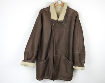 Op de afbeelding: Een bruine leren jas met een crème kleurige shearling voering en afwerking. De jas heeft een knoopsluiting, twee voorzakken en een reverskraag. De mouwen hebben shearling manchetten. De jas hangt aan een houten hanger.