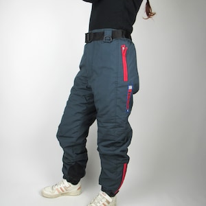 Op de afbeelding: Donkerblauwe ski broek met rode en blauwe accenten. De broek heeft een rits aan de zijkant en een zwarte riem.