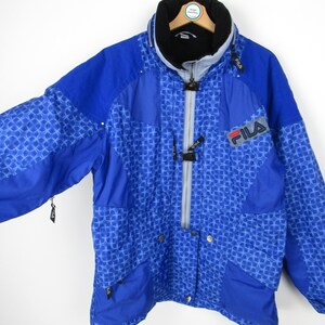 May include: A blue Fila jacket with a geometric pattern. The jacket has a high collar, a zipper closure, and the Fila logo on the chest. The jacket has multiple pockets and a black lining.