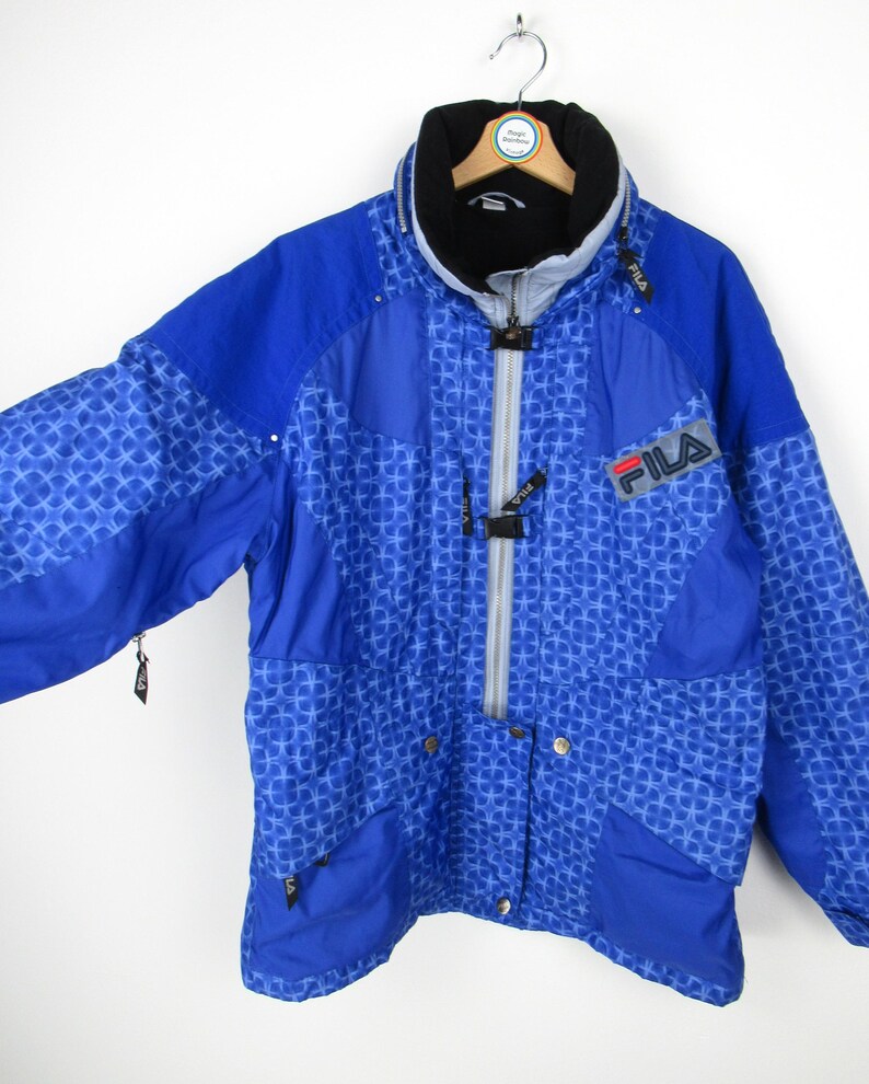 May include: A blue Fila jacket with a geometric pattern. The jacket has a high collar, a zipper closure, and the Fila logo on the chest. The jacket has multiple pockets and a black lining.