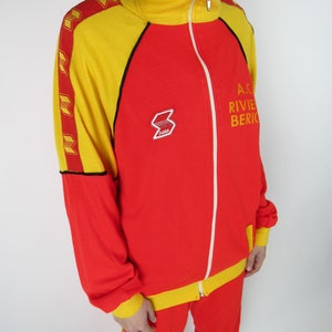 Puede incluir: Ch&aacute;ndal rojo y amarillo con cierre de cremallera y ribetes negros. La chaqueta tiene una raya amarilla en el lateral y los pantalones tienen una raya amarilla en la pierna. La chaqueta tiene un logotipo en el pecho izquierdo que dice "ARM". La chaqueta tambi&eacute;n tiene texto que dice "A.C. RIVIERA BERICO" en el lado derecho.