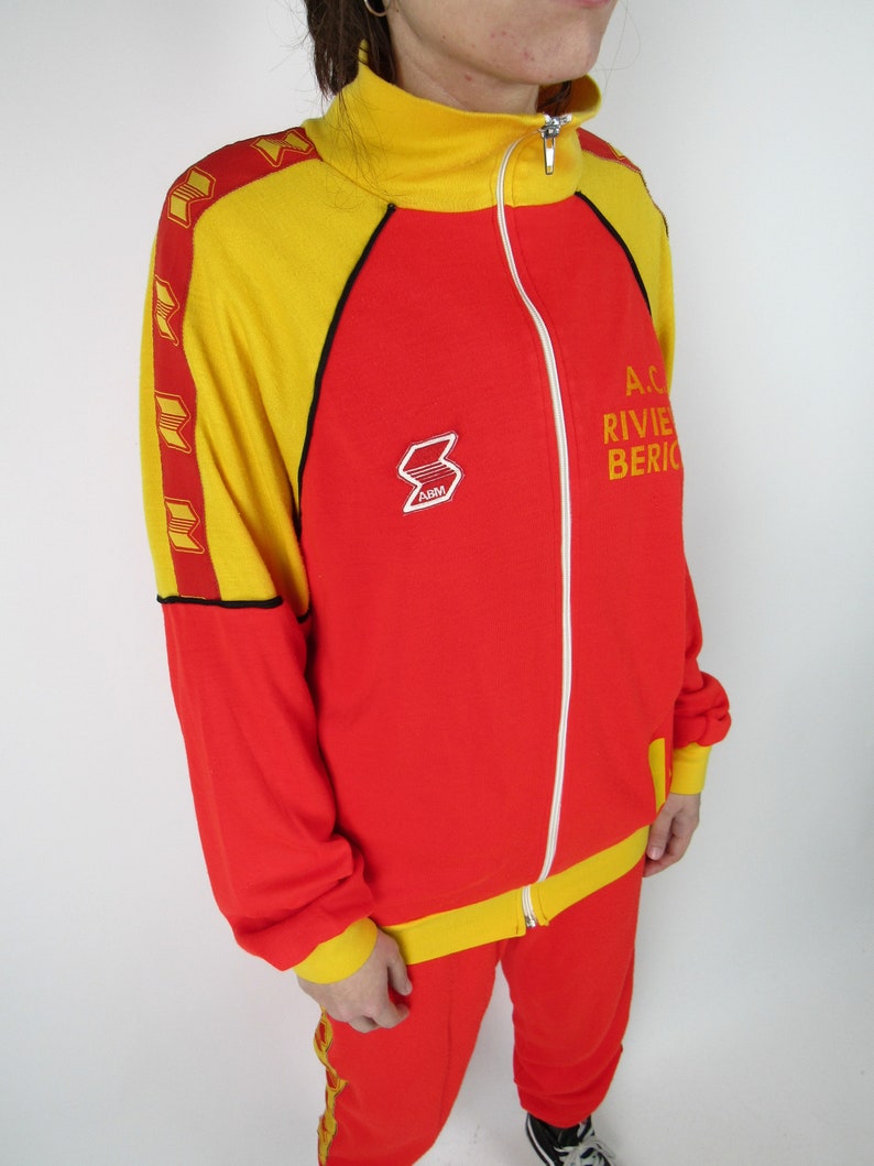 Puede incluir: Ch&aacute;ndal rojo y amarillo con cierre de cremallera y ribetes negros. La chaqueta tiene una raya amarilla en el lateral y los pantalones tienen una raya amarilla en la pierna. La chaqueta tiene un logotipo en el pecho izquierdo que dice "ARM". La chaqueta tambi&eacute;n tiene texto que dice "A.C. RIVIERA BERICO" en el lado derecho.