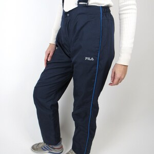 May include: Navy blue ski pants with a blue stripe down the side and the word "FILA" in white on the left leg. The pants have suspenders.