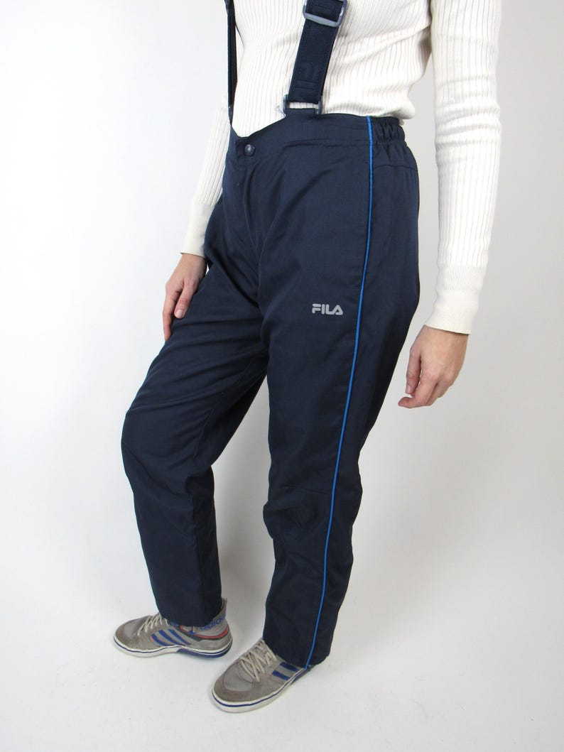 May include: Navy blue ski pants with a blue stripe down the side and the word "FILA" in white on the left leg. The pants have suspenders.