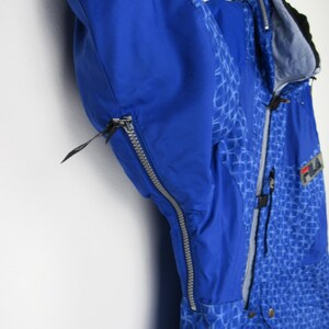 May include: A blue and patterned winter jacket with a zipper closure. The jacket has a hood and the brand name "FILA" is visible. The jacket is hanging on a hook against a white wall.