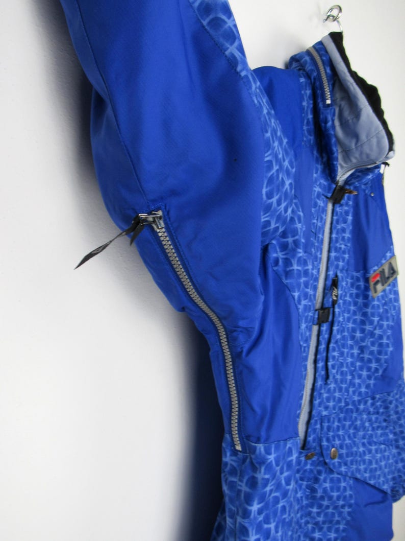 May include: A blue and patterned winter jacket with a zipper closure. The jacket has a hood and the brand name "FILA" is visible. The jacket is hanging on a hook against a white wall.