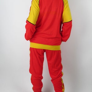 Puede incluir: Ch&aacute;ndal rojo y amarillo con ribetes negros. La chaqueta tiene cierre de cremallera y cuello alto. El pantal&oacute;n tiene pierna recta.