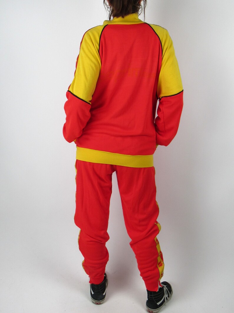 Puede incluir: Ch&aacute;ndal rojo y amarillo con ribetes negros. La chaqueta tiene cierre de cremallera y cuello alto. El pantal&oacute;n tiene pierna recta.