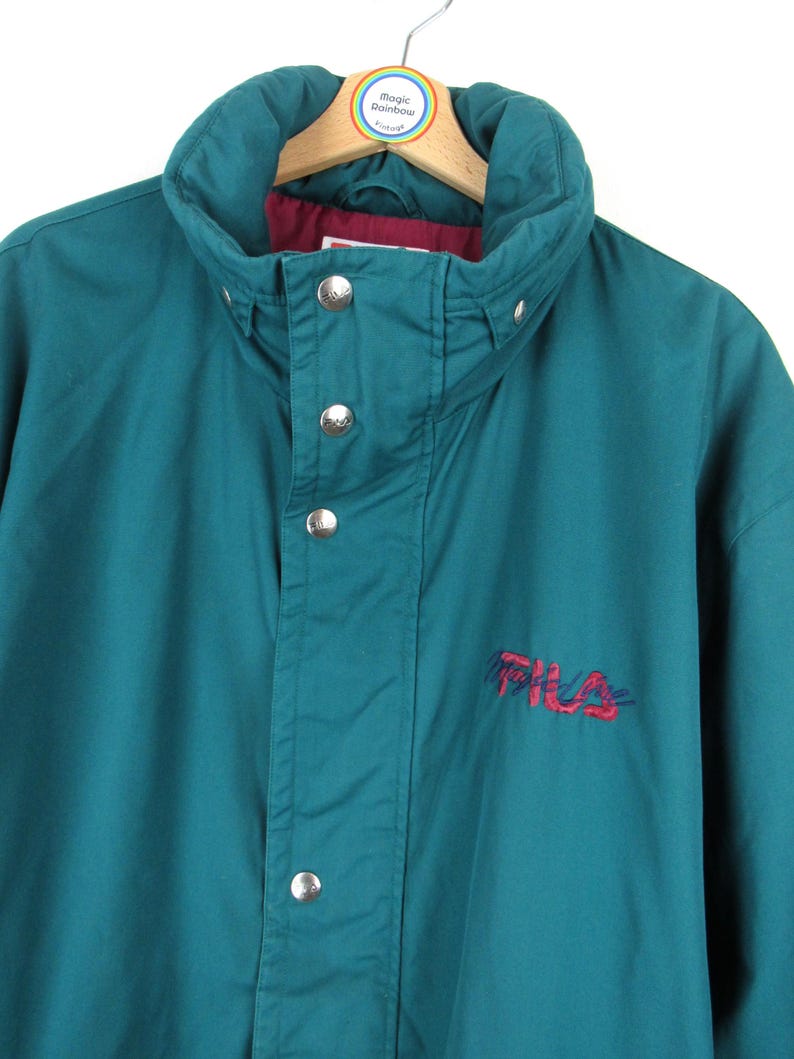 May include: Teal blue vintage Fila jacket with a red embroidered logo. The jacket has a high collar and a snap closure.