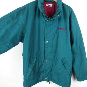 May include: A teal blue vintage Fila jacket with a red and white logo. The jacket has a zipper closure and a drawstring at the bottom.