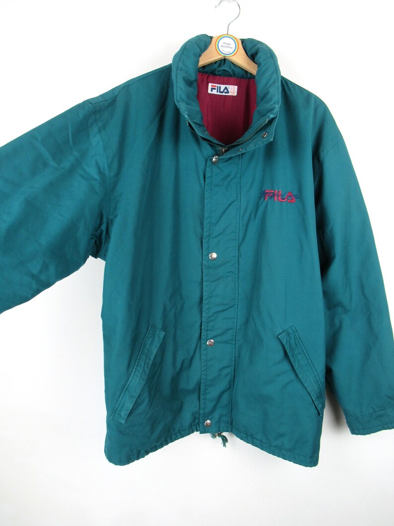 May include: A teal blue vintage Fila jacket with a red and white logo. The jacket has a zipper closure and a drawstring at the bottom.
