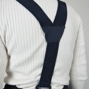 May include: A pair of dark blue suspenders with a white ribbed knit pattern. The suspenders are made of a woven fabric and have a clip closure.