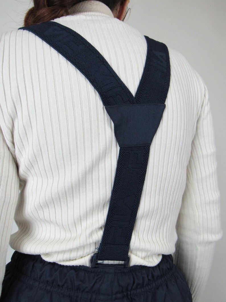 May include: A pair of dark blue suspenders with a white ribbed knit pattern. The suspenders are made of a woven fabric and have a clip closure.