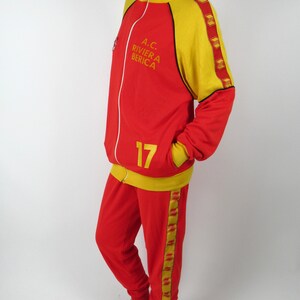Puede incluir: Ch&aacute;ndal rojo y amarillo con ribetes negros y una raya amarilla a lo largo del lateral del pantal&oacute;n. La chaqueta tiene cierre de cremallera y cuello alto. La chaqueta tiene el texto "A.C. RIVIERA BERICA" en la parte delantera. El pantal&oacute;n tiene el n&uacute;mero "17" en la parte delantera.