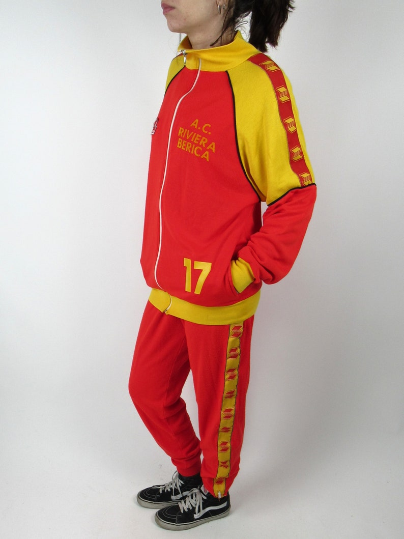 Puede incluir: Ch&aacute;ndal rojo y amarillo con ribetes negros y una raya amarilla a lo largo del lateral del pantal&oacute;n. La chaqueta tiene cierre de cremallera y cuello alto. La chaqueta tiene el texto "A.C. RIVIERA BERICA" en la parte delantera. El pantal&oacute;n tiene el n&uacute;mero "17" en la parte delantera.