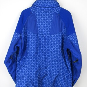 May include: A blue winter jacket with a geometric pattern. The jacket has a high collar, a zipper closure, and side pockets. The back of the jacket is visible, showing the intricate design. The jacket is hanging on a metal hanger.