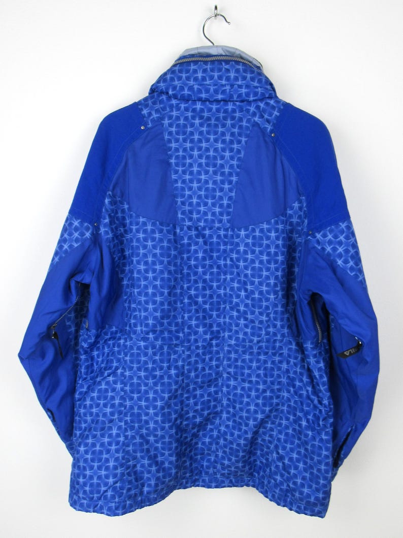 May include: A blue winter jacket with a geometric pattern. The jacket has a high collar, a zipper closure, and side pockets. The back of the jacket is visible, showing the intricate design. The jacket is hanging on a metal hanger.