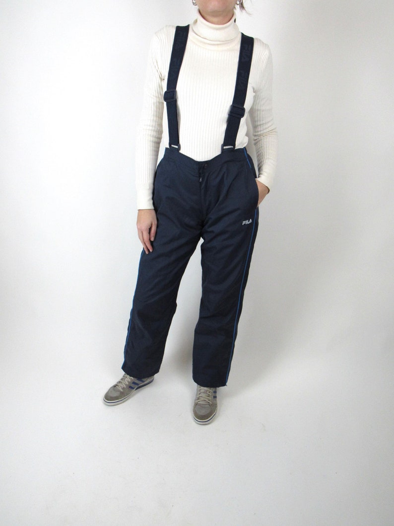 May include: Black ski pants with blue trim and suspenders. The pants have a drawstring waist and a Fila logo on the right leg.