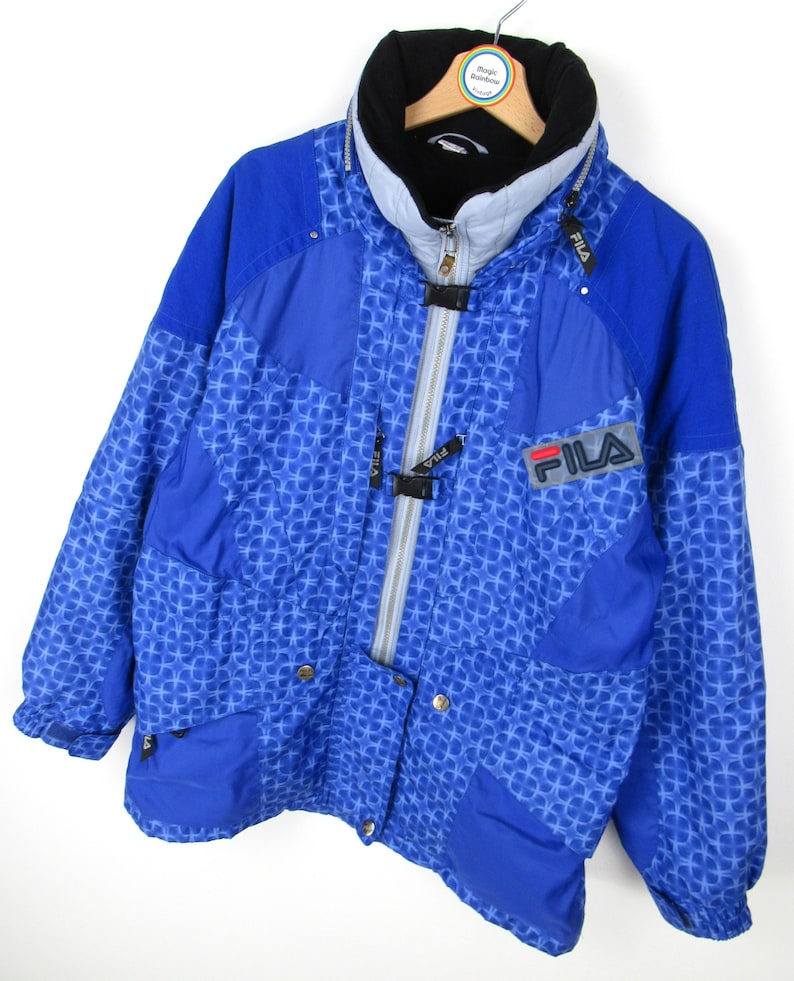 May include: A blue FILA winter jacket with a geometric pattern. The jacket features a high collar, a full-length zipper, and the FILA logo on the chest. The jacket has a black and gray color scheme.