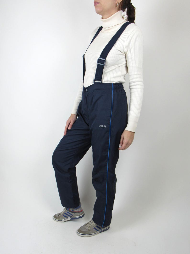May include: Navy blue ski pants with a blue stripe down the side and a FILA logo. The pants have suspenders and are made of a water-resistant material.