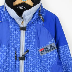 May include: A blue Fila ski jacket with a geometric pattern. The jacket features a black collar, a gray zipper, and the Fila logo on the chest. The jacket is hanging on a wooden hanger.