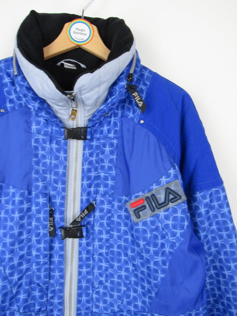 May include: A blue Fila ski jacket with a geometric pattern. The jacket features a black collar, a gray zipper, and the Fila logo on the chest. The jacket is hanging on a wooden hanger.