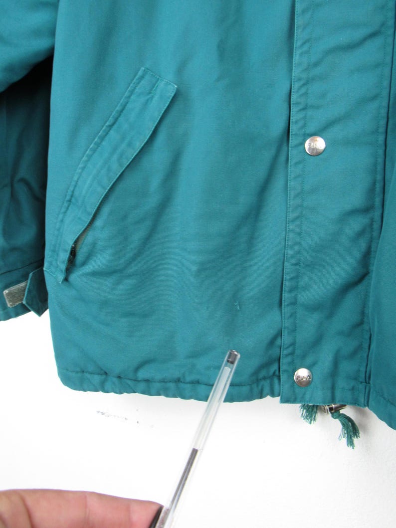 May include: Teal blue jacket with a zipper and button closure. The jacket has a pocket on the left side and a drawstring at the bottom.