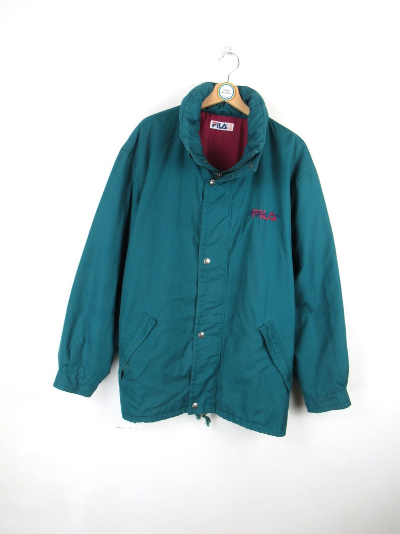 May include: A teal blue jacket with a red lining and a Fila logo on the front. The jacket has a zipper closure and a drawstring at the bottom.