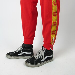 Puede incluir: Pantal&oacute;n de ch&aacute;ndal rojo con rayas laterales amarillas y rojas. El pantal&oacute;n de ch&aacute;ndal tiene una cremallera en la parte inferior de la pierna. La persona lleva zapatillas negras de ca&ntilde;a alta.