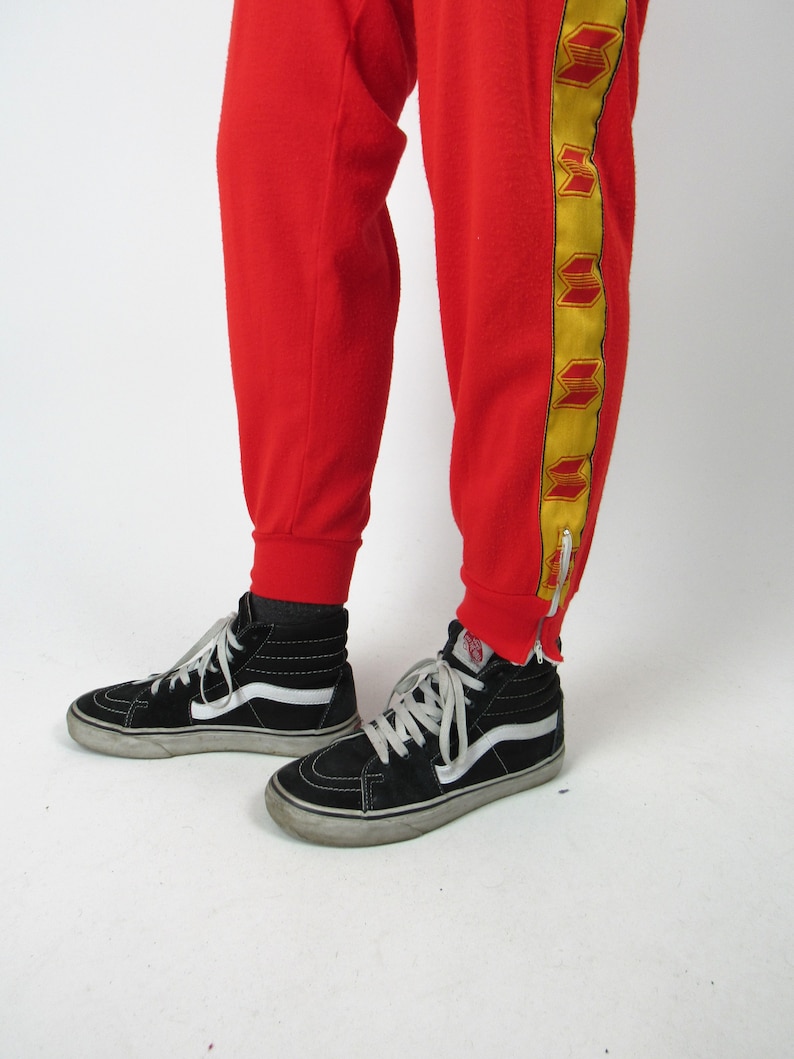 Puede incluir: Pantal&oacute;n de ch&aacute;ndal rojo con rayas laterales amarillas y rojas. El pantal&oacute;n de ch&aacute;ndal tiene una cremallera en la parte inferior de la pierna. La persona lleva zapatillas negras de ca&ntilde;a alta.